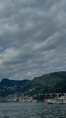 A scenic view featuring a mountainous landscape with dense greenery and a body of water in the foreground. The sky is mostly covered with thick, gray clouds. A boat with a Swiss flag is docked near the shore, and there are numerous small buildings scattered on the hillside.