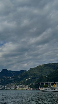 A scenic view featuring a mountainous landscape with dense greenery and a body of water in the foreground. The sky is mostly covered with thick, gray clouds. A boat with a Swiss flag is docked near the shore, and there are numerous small buildings scattered on the hillside.