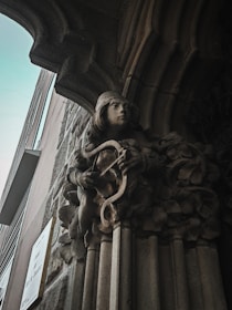 A stone sculpture depicting a serene figure playing a lyre is integrated into an ornate archway on a building facade. The craftsmanship is detailed, showing delicate features and flowing robes, surrounded by carved foliage. The surrounding architecture includes stone and metal elements, likely part of a historic or classical-style structure.
