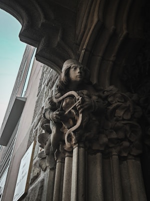 A stone sculpture depicting a serene figure playing a lyre is integrated into an ornate archway on a building facade. The craftsmanship is detailed, showing delicate features and flowing robes, surrounded by carved foliage. The surrounding architecture includes stone and metal elements, likely part of a historic or classical-style structure.