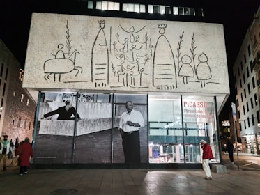 A building exterior featuring large, abstract line drawings resembling figures and a horse on a textured white surface. Beneath the artwork, there are large black-and-white photos of a person in an art studio setting. Several people are visible on the sidewalk near the building, with a modern cityscape in the background. The scene is illuminated by streetlights at night.