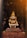 A weathered stone statue of a deity with a gold headdress and intricate carvings sits on a raised platform. The background shows a natural setting with dark, lush foliage, creating a mystical ambiance.
