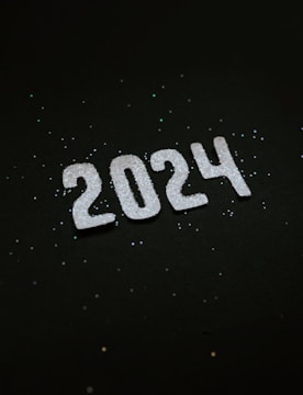 Large white numbers forming '2024' are placed against a dark background. The numbers are covered with a sparkly, glitter-like substance, and there are scattered specks of glitter surrounding the numbers.
