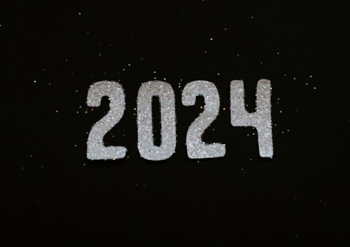 Glittery white numbers forming '2024' are prominently displayed against a dark background. Tiny specks of glitter are scattered around, creating a festive and celebratory vibe.