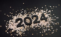 The image depicts the numbers 2024 in large, bold black digits placed on a dark surface. Surrounding the numbers are numerous small star-shaped confetti pieces in metallic gold, silver, and white colors.