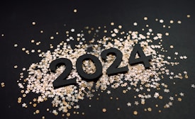 The image depicts the numbers 2024 in large, bold black digits placed on a dark surface. Surrounding the numbers are numerous small star-shaped confetti pieces in metallic gold, silver, and white colors.