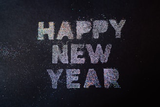 Bright and colorful glitter letters spell out 'Happy New Year' on a dark background, with scattered glitter particles surrounding the text.