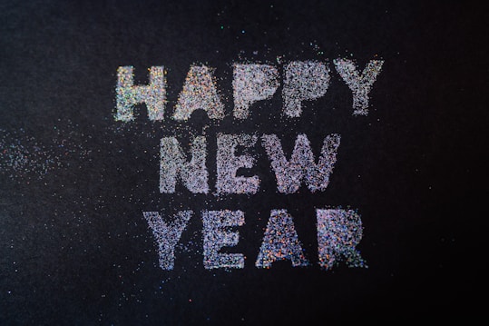 Bright and colorful glitter letters spell out 'Happy New Year' on a dark background, with scattered glitter particles surrounding the text.