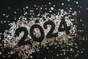 The image features black numerals forming the year 2024 surrounded by scattered star-shaped confetti. The confetti varies in color, including shades of silver and gold, creating a celebratory atmosphere.