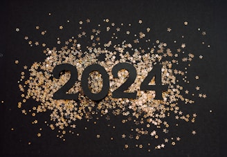 The number 2024 is prominently displayed in bold black against a dark background. Surrounding the numbers are small, shiny gold and silver star-shaped confetti that create a festive atmosphere.
