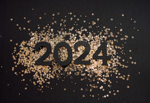 The number 2024 is prominently displayed in bold black against a dark background. Surrounding the numbers are small, shiny gold and silver star-shaped confetti that create a festive atmosphere.