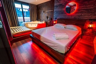 A cozy bedroom featuring a large wooden bed with white linens and a neatly placed towel. The room is illuminated by warm red lights, giving it a romantic and soothing ambiance. Beside the bed, there is a Jacuzzi tub with a luxurious appearance, and a lounge chair. Dark curtains cover the windows, adding to the intimate atmosphere. The walls are decorated with wood panels and a circular mirror is highlighted by red lighting.