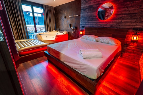 A cozy bedroom featuring a large wooden bed with white linens and a neatly placed towel. The room is illuminated by warm red lights, giving it a romantic and soothing ambiance. Beside the bed, there is a Jacuzzi tub with a luxurious appearance, and a lounge chair. Dark curtains cover the windows, adding to the intimate atmosphere. The walls are decorated with wood panels and a circular mirror is highlighted by red lighting.