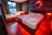 A cozy bedroom featuring a large wooden bed with white linens and a neatly placed towel. The room is illuminated by warm red lights, giving it a romantic and soothing ambiance. Beside the bed, there is a Jacuzzi tub with a luxurious appearance, and a lounge chair. Dark curtains cover the windows, adding to the intimate atmosphere. The walls are decorated with wood panels and a circular mirror is highlighted by red lighting.