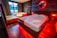 A cozy bedroom featuring a large wooden bed with white linens and a neatly placed towel. The room is illuminated by warm red lights, giving it a romantic and soothing ambiance. Beside the bed, there is a Jacuzzi tub with a luxurious appearance, and a lounge chair. Dark curtains cover the windows, adding to the intimate atmosphere. The walls are decorated with wood panels and a circular mirror is highlighted by red lighting.