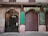 A street view of a building facade featuring intricate iron gates and an old-fashioned storefront with a green and gold sign that reads 'Farmacia.' The gate on the left side is intricately designed, while the storefront gate on the right is closed and painted with graffiti. Above the gates are decorative architectural details, and there are balconies with closed shutters. A tree branch extends into the frame from the right side.
