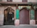 A street view of a building facade featuring intricate iron gates and an old-fashioned storefront with a green and gold sign that reads 'Farmacia.' The gate on the left side is intricately designed, while the storefront gate on the right is closed and painted with graffiti. Above the gates are decorative architectural details, and there are balconies with closed shutters. A tree branch extends into the frame from the right side.
