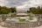A grand villa with symmetrical architecture sits in the distance, bordered by lush green trees with autumnal hues. A long reflecting pool stretches in front of the villa, with statues on either side and water lilies floating on the surface. The sky is overcast, casting a serene and contemplative atmosphere.