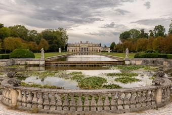 A grand villa with symmetrical architecture sits in the distance, bordered by lush green trees with autumnal hues. A long reflecting pool stretches in front of the villa, with statues on either side and water lilies floating on the surface. The sky is overcast, casting a serene and contemplative atmosphere.