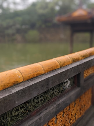 A decorative stone wall featuring intricate patterns with earthy tones dominates the foreground. The top of the wall has a rounded, oxidized orange finish, and beneath are ornate green and brown tiles. In the background, a blurred out-of-focus scene suggests a natural setting with lush greenery and a traditional structure.