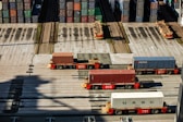 Several automated cargo trucks carrying shipping containers are lined up on a concrete port terminal. Numerous colorful stacked containers are visible in the background, suggesting a busy port. The ground is marked with shadows and tire tracks.