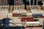 Several automated cargo trucks carrying shipping containers are lined up on a concrete port terminal. Numerous colorful stacked containers are visible in the background, suggesting a busy port. The ground is marked with shadows and tire tracks.