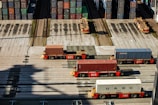 Several automated cargo trucks carrying shipping containers are lined up on a concrete port terminal. Numerous colorful stacked containers are visible in the background, suggesting a busy port. The ground is marked with shadows and tire tracks.