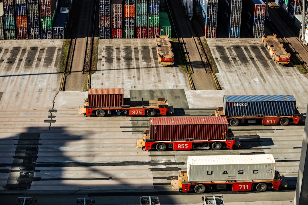 a group of trucks parked next to each other in a parking lot, Aerial view of a container terminal with several trucks transporting colorful cargo containers, casting long shadows in the bright sunlight, indicative of bustling port activity.
