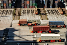 Several automated cargo trucks carrying shipping containers are lined up on a concrete port terminal. Numerous colorful stacked containers are visible in the background, suggesting a busy port. The ground is marked with shadows and tire tracks.