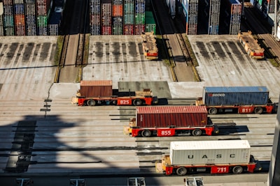 Several automated cargo trucks carrying shipping containers are lined up on a concrete port terminal. Numerous colorful stacked containers are visible in the background, suggesting a busy port. The ground is marked with shadows and tire tracks.