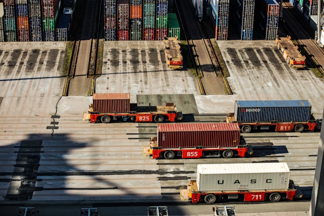 Several automated cargo trucks carrying shipping containers are lined up on a concrete port terminal. Numerous colorful stacked containers are visible in the background, suggesting a busy port. The ground is marked with shadows and tire tracks.