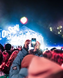 Crowd of people gathered at an outdoor concert at night, with one person holding up a smartphone to capture the performance. Bright stage lights illuminate the scene with vibrant colors, creating an energetic atmosphere. The stage features the word 'LOUSSEAU' in large, illuminated letters.