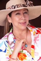 A person wearing a straw hat with a leopard print ribbon, gold hoop earrings, and a colorful floral-patterned shirt. The individual is smiling gently and has a gold chain necklace with a pendant featuring an eye symbol. There is a bracelet on the left wrist.