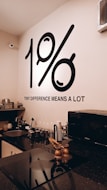 A modern kitchen setup with neatly arranged utensils and appliances on a black countertop. The wall features a prominent black graphic displaying '1%' along with the phrase 'TINY DIFFERENCE MEANS A LOT'. The environment appears organized and minimalistic with a microwave, various kitchen tools, and a sink visible.