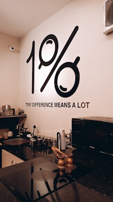 A modern kitchen setup with neatly arranged utensils and appliances on a black countertop. The wall features a prominent black graphic displaying '1%' along with the phrase 'TINY DIFFERENCE MEANS A LOT'. The environment appears organized and minimalistic with a microwave, various kitchen tools, and a sink visible.