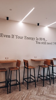 A modern interior space with high wooden stools and tables attached to the wall. A motivational quote is written on the wall: 'Even If Your Energy Is 99%... You still need 1%'. The floor is tiled in a light wood pattern, and linear ceiling lights are present.