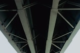 A striking view of the underside of a large bridge with metal beams and crossbars. The structure appears robust and industrial, showcasing the engineering and design of modern infrastructure. The perspective is upward, highlighting the geometric patterns formed by the intersecting beams.