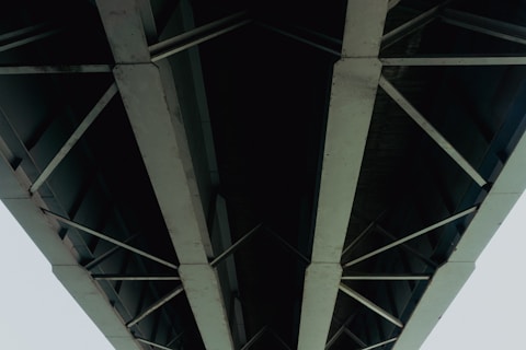 A striking view of the underside of a large bridge with metal beams and crossbars. The structure appears robust and industrial, showcasing the engineering and design of modern infrastructure. The perspective is upward, highlighting the geometric patterns formed by the intersecting beams.