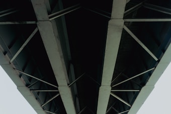 A striking view of the underside of a large bridge with metal beams and crossbars. The structure appears robust and industrial, showcasing the engineering and design of modern infrastructure. The perspective is upward, highlighting the geometric patterns formed by the intersecting beams.