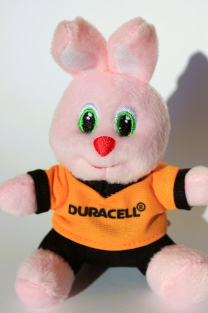 A plush pink bunny with large, shiny green eyes and a red nose. It wears a bright orange and black shirt with a logo on it.