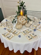 A round table covered with a white cloth is arranged with small craft kits, each containing a card with line art drawings and a watercolor paint set. A decorative sign featuring the phrase 'give thanks' is placed on the table. Beside it, small decorative pumpkins and a lantern with a candle are enhanced by intertwined green vines.