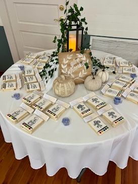 A round table covered with a white cloth is arranged with small craft kits, each containing a card with line art drawings and a watercolor paint set. A decorative sign featuring the phrase 'give thanks' is placed on the table. Beside it, small decorative pumpkins and a lantern with a candle are enhanced by intertwined green vines.
