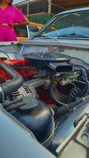 A detailed view of a car engine with various components such as hoses, wires, and metallic parts. In the background, a person wearing a bright pink outfit stands next to a car with an orange exterior and brown interior. The setting appears to be an outdoor area with a chain-link fence covered in green foliage.