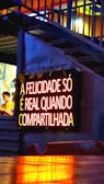 A glowing neon sign with the text 'A FELICIDADE SÓ É REAL QUANDO COMPARTILHADA' is mounted on a wall. The lighting gives a warm, inviting feel, and the background features some stairs with accent lighting. A person is partially visible to the side, blurred and possibly moving.