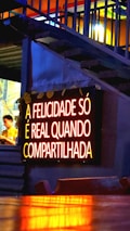 A glowing neon sign with the text 'A FELICIDADE S&Oacute; &Eacute; REAL QUANDO COMPARTILHADA' is mounted on a wall. The lighting gives a warm, inviting feel, and the background features some stairs with accent lighting. A person is partially visible to the side, blurred and possibly moving.