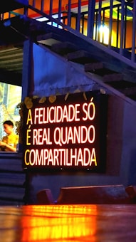 A glowing neon sign with the text 'A FELICIDADE SÓ É REAL QUANDO COMPARTILHADA' is mounted on a wall. The lighting gives a warm, inviting feel, and the background features some stairs with accent lighting. A person is partially visible to the side, blurred and possibly moving.