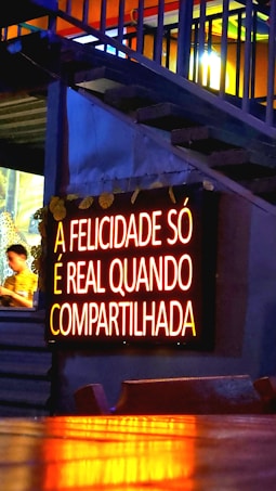 A glowing neon sign with the text 'A FELICIDADE SÓ É REAL QUANDO COMPARTILHADA' is mounted on a wall. The lighting gives a warm, inviting feel, and the background features some stairs with accent lighting. A person is partially visible to the side, blurred and possibly moving.