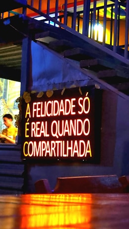 A glowing neon sign with the text 'A FELICIDADE S&Oacute; &Eacute; REAL QUANDO COMPARTILHADA' is mounted on a wall. The lighting gives a warm, inviting feel, and the background features some stairs with accent lighting. A person is partially visible to the side, blurred and possibly moving.