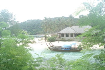 A serene beach setting with lush greenery in the foreground. A traditional wooden boat with a blue roof floats on clear turquoise waters. In the background, a thatched-roof building is nestled among tall palm trees, and a dense green hillside rises in the distance.