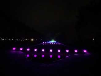 A series of glowing orbs arranged in a symmetrical pattern creating a pathway that appears to extend into the distance. The orbs are primarily purple, with a few shifting into pink and blue tones as they recede. The scene is set against a dark background, emphasizing the luminous quality of the lights.